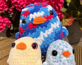 May include: Three crocheted bird toys. The largest bird is blue, red, and white with a bright orange beak. The smaller birds are yellow and light green with orange beaks. The text "Bertie the Bird" is visible in the background.
