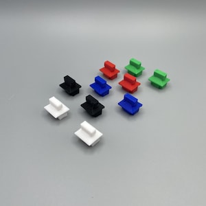 Może przedstawiać: Zestaw małych plastikowych elementów w różnych kolorach, w tym białym, czarnym, niebieskim, czerwonym i zielonym. Elementy mają kształt małych kwadratów z podniesionym wierzchołkiem.