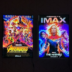 May include: Two framed movie posters. The left poster is for "Avengers: Infinity War" with a cast of characters and the date April 27. The right poster is for "The Marvels" with the text "Experience it in IMAX" and the date November 10.