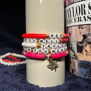 May include: Stack of beaded bracelets in red, white, and pink with gold accents. The bracelets spell out "RED", "TOO WELL", "TAYLOR", and "NEW YOU WERE". A gold maple leaf charm hangs from one bracelet. A smaller bracelet is visible in the background.