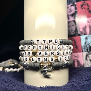May include: A set of grey and white beaded bracelets with silver and gold accents. The bracelets spell out the words "TTPD", "FORTNIGHT", "ITS", "VERSION", and "SO LONG". A silver bird charm hangs from the bottom of the bracelets.
