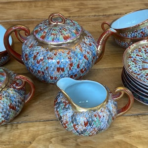 Puede incluir: Un juego de té vintage con un patrón moteado azul, rojo y dorado. El juego incluye una tetera, azucarero, lechera, tazas de té y platillos. Los bordes y las asas son de color marrón rojizo.