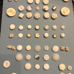 May include: Assortment of vintage buttons in various shapes, sizes, and colors. The buttons include floral, geometric, and textured designs. Some buttons are white, cream, and beige, while others have iridescent or metallic accents. The buttons are arranged on a blue surface.