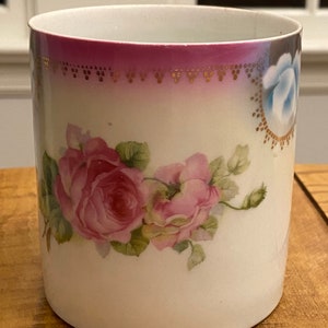 May include: A white ceramic mug with a pink and gold floral design. The mug features pink roses and blue flowers with gold accents.
