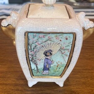 May include: A small, off-white ceramic container with a lid and a decorative handle. The container features a hand-painted scene of a person with an umbrella under a cherry blossom tree. The container has a gold trim and is supported by three small feet.