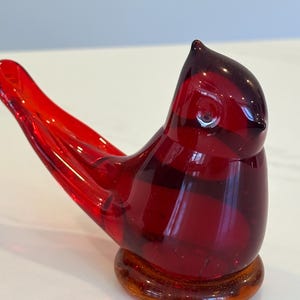 Könnte beinhalten: Eine leuchtend rote Glasvogel-Figur mit glänzender Oberfläche. Der Vogel sitzt auf einem kreisförmigen, bernsteinfarbenen Sockel. Das Design der Figur zeigt einen langen Schwanz und einen gebogenen Schnabel, was eine detaillierte und künstlerische Darstellung bietet.