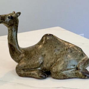 Könnte beinhalten: Eine Kamelfigur aus Keramik in sitzender Position mit einer glänzenden, olivgrünen Sprenkelglasur. Die Skulptur zeigt einen langen Hals, einen einzelnen Höcker und angewinkelte Beine mit einer glatten, reflektierenden Oberfläche.