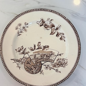 May include: A round, off-white plate with a brown floral and landscape design. The plate features a detailed border and a central scene of figures and animals. The design includes flowers and foliage, creating a vintage aesthetic.