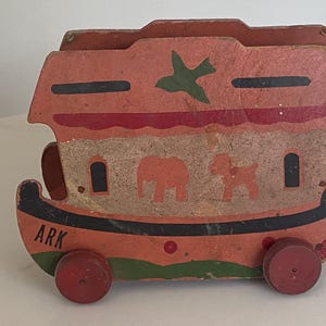 May include: Vintage wooden toy ark painted in shades of orange, red, and green. The ark features a green bird, an elephant, and a dog. The word "ARK" is painted on the side. The toy has red wheels.