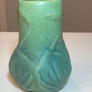 May include: A small, turquoise-colored ceramic vase with a bulbous base and a cylindrical top. The vase features a crackle glaze and stylized leaf-like patterns. The color transitions from a lighter green at the top to a deeper teal at the base.