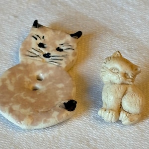 Puede incluir: Dos figuras de gato de cerámica. Una es un gato en forma de botón con un diseño veteado rosa claro y blanco, orejas negras y bigotes. La otra es una pequeña figura de gato blanquecino en posición sentada.