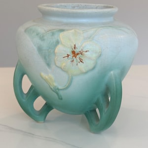 May include: A light blue ceramic vase with a floral design. The vase has a rounded body and three curved legs. A white flower with brown accents is embossed on the front. The vase has a crackled glaze finish.