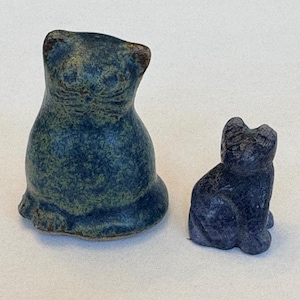 Puede incluir: Dos figuras de gato de cerámica. Una es un gato sentado más grande en tonos azules y verdes. La otra es un gato sentado más pequeño en azul oscuro. Ambos tienen un aspecto texturizado y están sobre una superficie blanca.