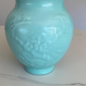 May include: A light blue ceramic vase with a raised floral design. The vase has a rounded body and a wide mouth.