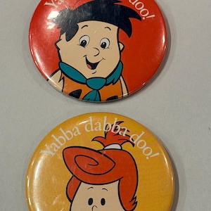 Puede incluir: Dos botones redondos con personajes de la serie de dibujos animados Los Picapiedra. Un botón tiene a Pedro Picapiedra con la frase "Yabba Dabba Doo!" en texto blanco sobre un fondo rojo. El otro botón tiene a Vilma Picapiedra con la misma frase sobre un fondo amarillo.