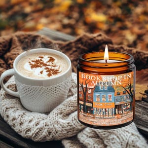 May include: A lit candle in a glass jar with a label that reads "Book Shop in Autumn" next to a white mug of coffee with a decorative design on top. The mug is wrapped in a white knitted cozy. The background is blurred with fall leaves.