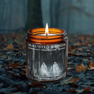 May include: A lit, amber-colored glass jar candle with the text "HAUNTED WOODS" and a forest scene with ghosts. The candle is surrounded by fallen leaves, creating a spooky atmosphere.