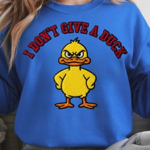 May include: Blue sweatshirt with the embroidered phrase "I DON'T GIVE A DUCK" in red. The design features a yellow cartoon duck with an angry expression. The duck has orange feet and a beak.