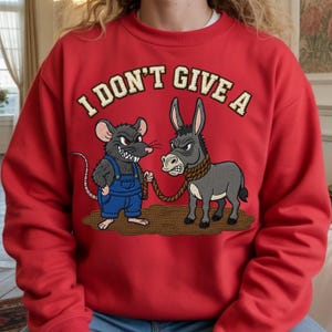 May include: Red sweatshirt with the text "I DON'T GIVE A" above a cartoon illustration of a gray rat in blue overalls holding a rope attached to a gray donkey. The donkey has a rope around its neck.
