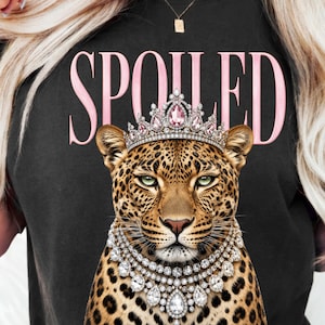 May include: Black t-shirt with the word "SPOILED" in pink lettering. The shirt features a leopard illustration wearing a jeweled crown and necklace. The leopard has green eyes and a spotted coat.