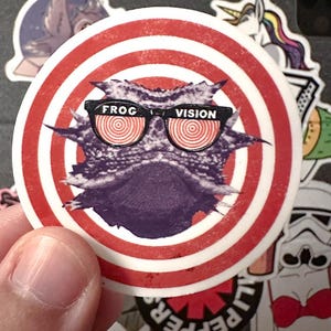 Może przedstawiać: Okrągła naklejka z fioletową jaszczurką rogatą w czarnych okularach przeciwsłonecznych z napisem "FROG VISION". Tło ma czerwono-białe okręgi koncentryczne. Inne naklejki są widoczne w tle.