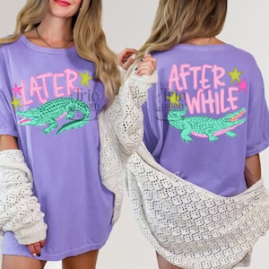 May include: A lilac-colored t-shirt featuring a green alligator graphic with the text "LATER" on the front and "AFTER WHILE" on the back. The design includes pink and yellow star accents, creating a playful and casual look.