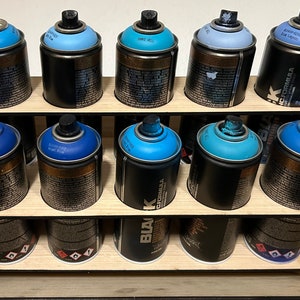 Puede incluir: Una estantería de madera con 12 botes de pintura en aerosol en varios tonos de azul. Los botes están dispuestos en dos filas de seis. La estantería está hecha de madera y tiene un acabado marrón oscuro.