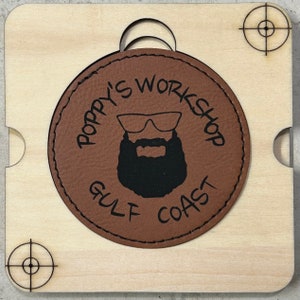 May include: A wooden square with a circular brown leather patch. The patch features the text "POPPY'S WORKSHOP GULF COAST" around a graphic of a bearded man wearing sunglasses. Target-like markings are in each corner.