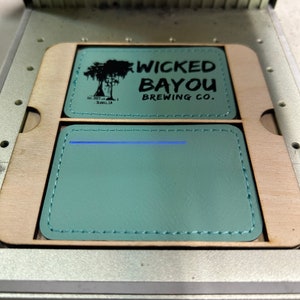 May include: Two teal leather coasters with stitched edges are visible inside a laser engraving machine. The top coaster has the text "WICKED BAYOU BREWING CO." and a tree graphic. The bottom coaster is blank.