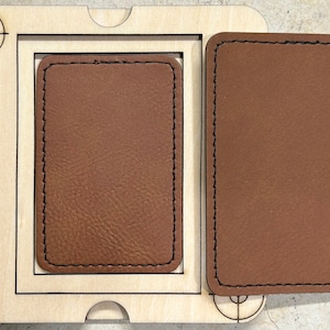 May include: A wooden jig with a rectangular cutout and a separate brown leather piece with black stitching. Another brown leather piece is next to the jig. The jig has a target design on the corner.
