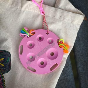 May include: A pink plastic organizer with six circular holes for holding small items. The organizer has a chain attached to it and is hanging from a white tote bag. The text "POPPY'S WORKSHOP GC" is printed on the organizer.