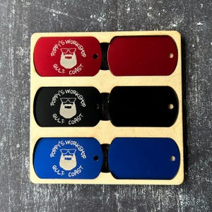 May include: Three metal dog tags in a wooden display stand. The top tag is red, the middle tag is black, and the bottom tag is blue. Each tag has a white engraved design of a bearded man wearing sunglasses and the text "Poppy's Workshop Gulf Coast".