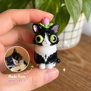 Op de afbeelding: Een handgemaakt zwart-wit kattenbeeldje met grote groene ogen en een roze bloem op het hoofd. Het beeldje wordt in de hand van een persoon gehouden. De tekst "Made from your cat" staat ook in de afbeelding.