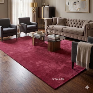 May include: A large rectangular deep red area rug with a soft texture, positioned in a living room setting featuring a tufted beige sofa, three dark gray armchairs, a glass-topped coffee table, and the text heritage by fiza in the corner.