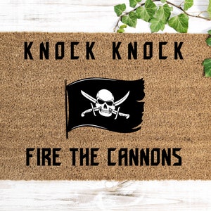 May include: A brown doormat with the text "Knock Knock" and "Fire the Cannons" in black. A black pirate flag with a white skull and crossbones is in the center.