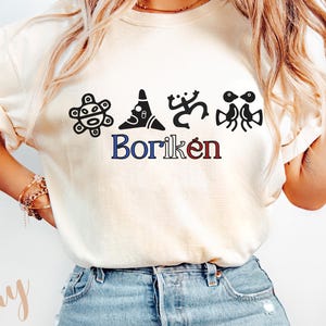 May include: Ivory t-shirt with black graphic designs and the word "Borik&eacute;n" in blue and red. The shirt has short sleeves and is worn with blue denim jeans. The style is casual and comfortable, suitable for everyday wear.