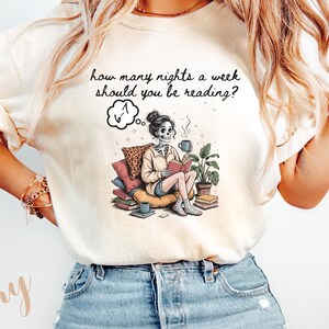 May include: Ivory-colored t-shirt featuring a whimsical illustration of a skeleton reading a book, with a coffee cup and plants. The text above the illustration reads, "how many nights a week should you be reading?" The skeleton is seated on pillows.