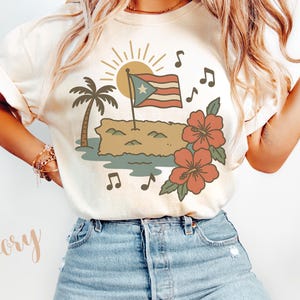 Op de afbeelding: Ivoor kleurig T-shirt met een grafisch ontwerp met een Puerto Ricaanse vlag, eiland, palmboom, zon, muzieknoten en hibiscusbloemen. Het ontwerp is in bruin, groen, rood en blauw. Het woord "Ivory" is in een cursief lettertype geschreven.