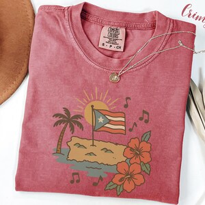 Op de afbeelding: K малиновое T-shirt met een afbeelding van de Puerto Ricaanse vlag, een palmboom en hibiscusbloemen. Het ontwerp bevat muzieknoten en een zon. Een zilveren ketting met een hanger ligt op het shirt.