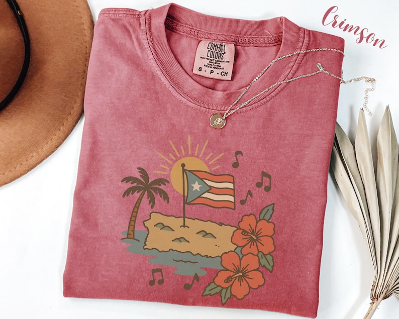 Op de afbeelding: K малиновое T-shirt met een afbeelding van de Puerto Ricaanse vlag, een palmboom en hibiscusbloemen. Het ontwerp bevat muzieknoten en een zon. Een zilveren ketting met een hanger ligt op het shirt.