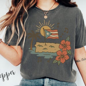 Op de afbeelding: Donkergrijs t-shirt met een grafische afbeelding van de Puerto Ricaanse vlag, een palmboom, zon en hibiscusbloemen. Het ontwerp bevat muzieknoten en een afbeelding van het eiland. Het woord "Pepper" is cursief geschreven.