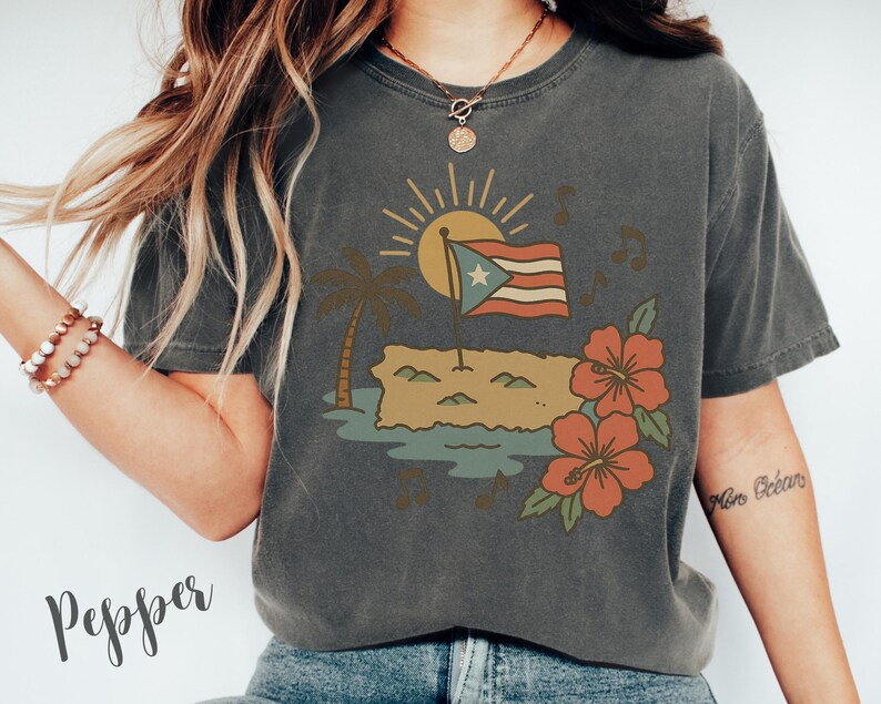 Op de afbeelding: Donkergrijs t-shirt met een grafische afbeelding van de Puerto Ricaanse vlag, een palmboom, zon en hibiscusbloemen. Het ontwerp bevat muzieknoten en een afbeelding van het eiland. Het woord "Pepper" is cursief geschreven.