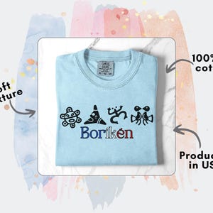 May include: Pale blue crew neck t-shirt with black graphic designs and the word "Borik&eacute;n" in blue and red. The shirt is made of 100% US cotton.