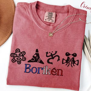 May include: Crimson-coloured t-shirt with a crew neck and the word "Borik&eacute;n" in blue and red. The shirt features black graphic designs and a Comfort Colors tag. A silver necklace with a pendant rests on the shirt.