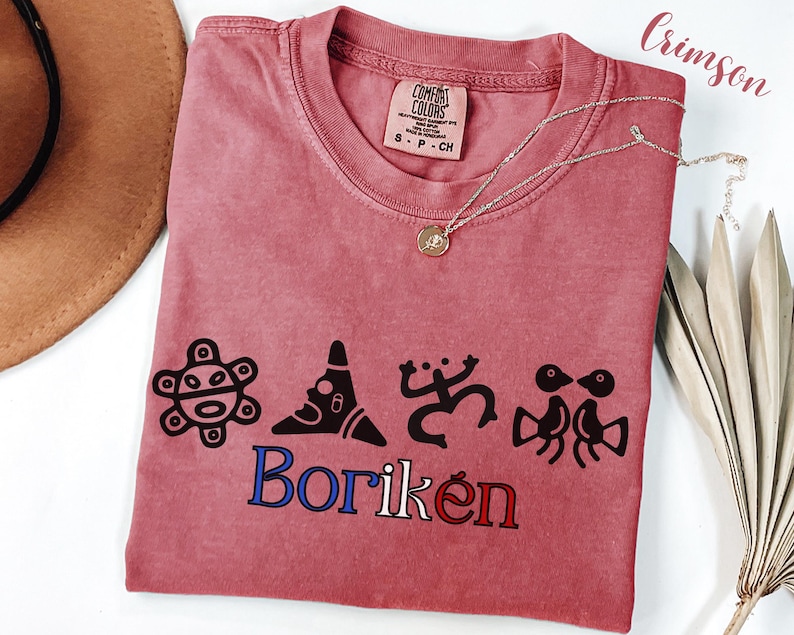 May include: Crimson-coloured t-shirt with a crew neck and the word "Borik&eacute;n" in blue and red. The shirt features black graphic designs and a Comfort Colors tag. A silver necklace with a pendant rests on the shirt.
