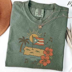 Op de afbeelding: Een mosgroen t-shirt met een afbeelding van de Puerto Ricaanse vlag, een palmboom en hibiscusbloemen. Het shirt heeft een ronde hals en korte mouwen. Het merklabel "Comfort Colors" is zichtbaar.