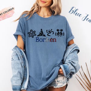 May include: A blue jean coloured t-shirt featuring the word "Borik&eacute;n" in blue and red, with black graphic designs above it. The shirt has short sleeves and a relaxed fit. The words "Comfort Colors" are in a pink banner.