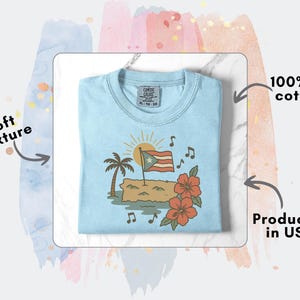 Op de afbeelding: Lichtblauw T-shirt met een afbeelding van de Puerto Ricaanse vlag, een palmboom, bloemen en muzieknoten. Het shirt is gemaakt van 100% US katoen en heeft een zachte textuur. Geproduceerd in de VS.