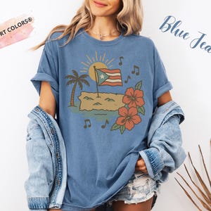 Op de afbeelding: Blauw spijkerbroekkleurig T-shirt met een afbeelding van een Puerto Ricaanse vlag, palmboom, zon, muzieknoten en hibiscusbloemen. De tekst "Blue Jean" is cursief geschreven. Het shirt heeft de tekst "Comfort Colors&reg;" linksboven.