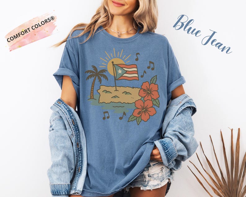 Op de afbeelding: Blauw spijkerbroekkleurig T-shirt met een afbeelding van een Puerto Ricaanse vlag, palmboom, zon, muzieknoten en hibiscusbloemen. De tekst "Blue Jean" is cursief geschreven. Het shirt heeft de tekst "Comfort Colors&reg;" linksboven.